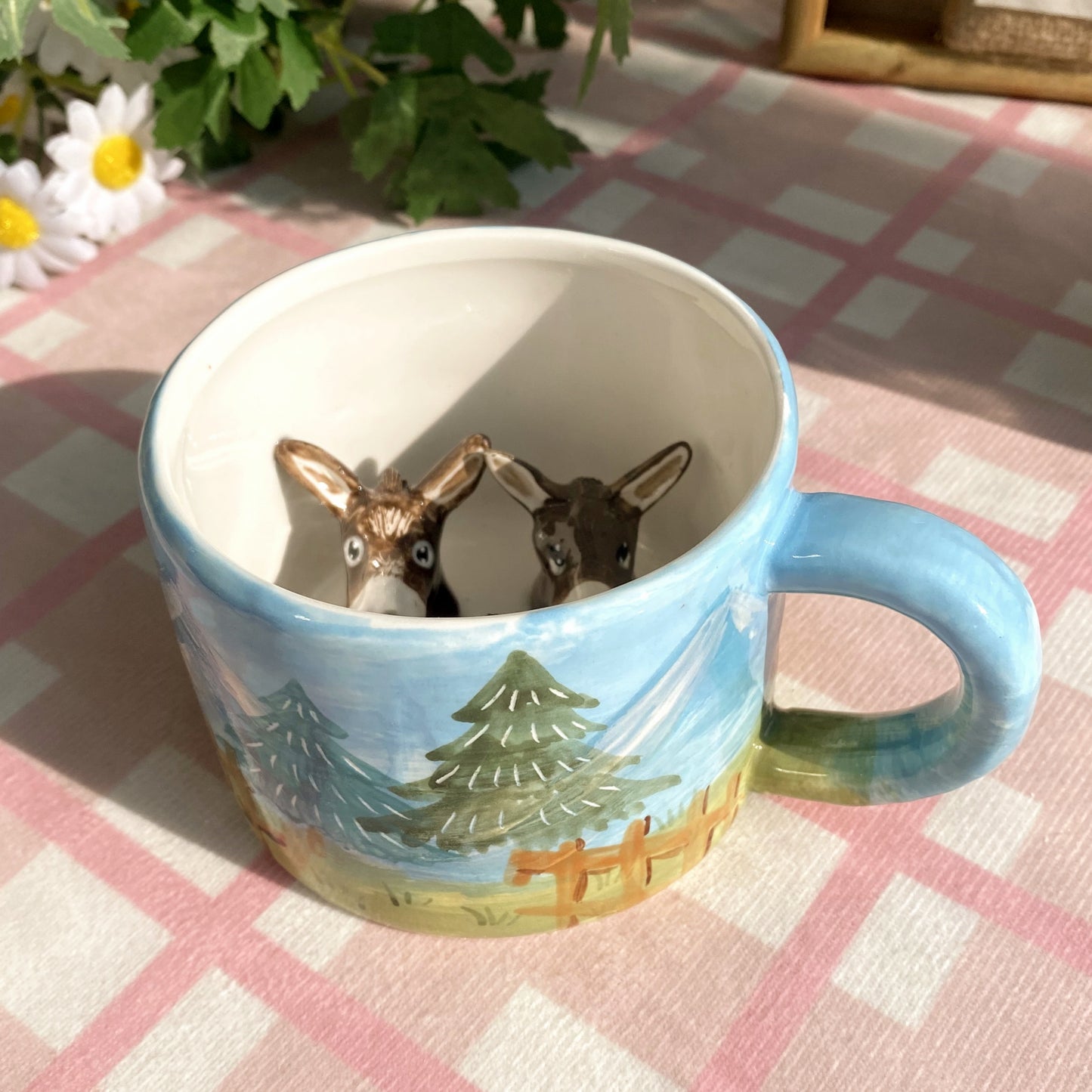 Hand-painted ceramic mug with blue handle and scenic forest landscape, featuring two 3D donkey figures inside the cup