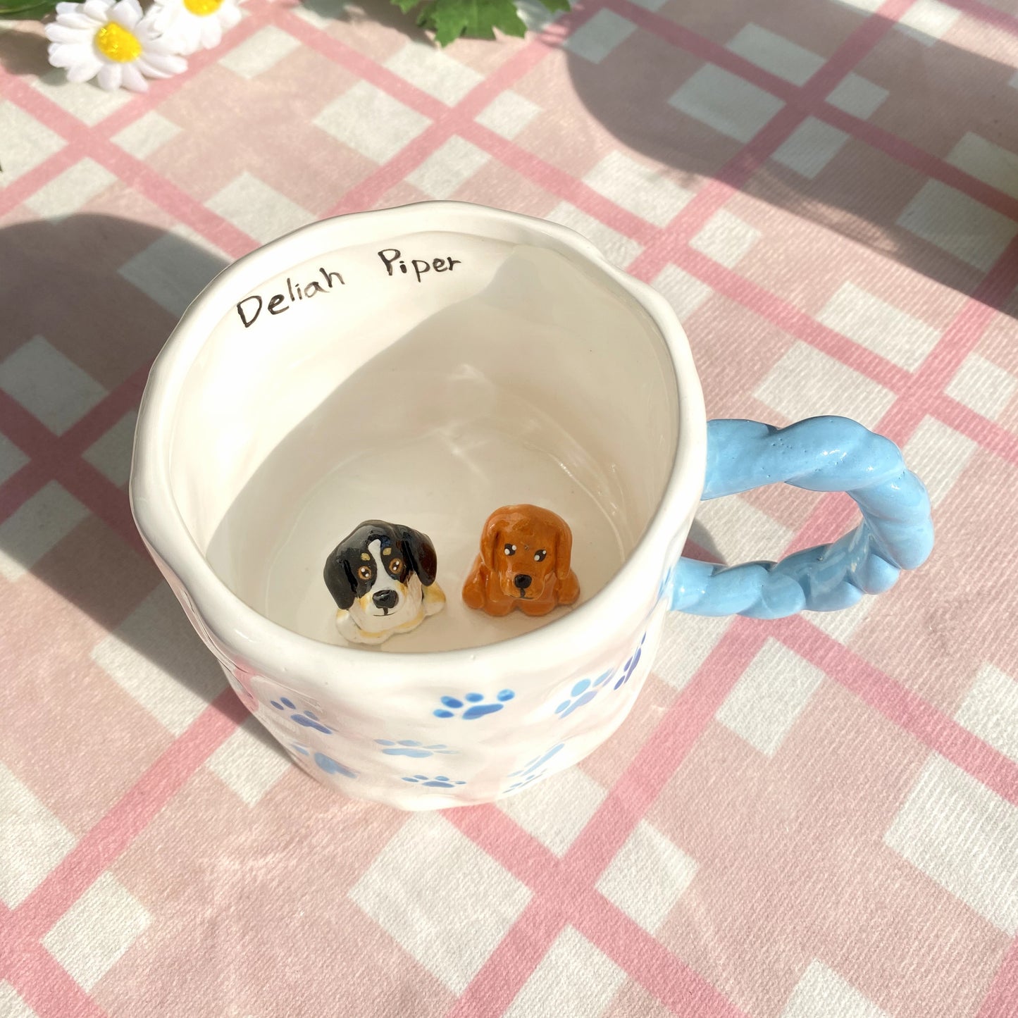 White ceramic mug with braided blue handle and paw prints, featuring two 3D puppy heads inside and the names "Deliah Piper" handwritten on the rim