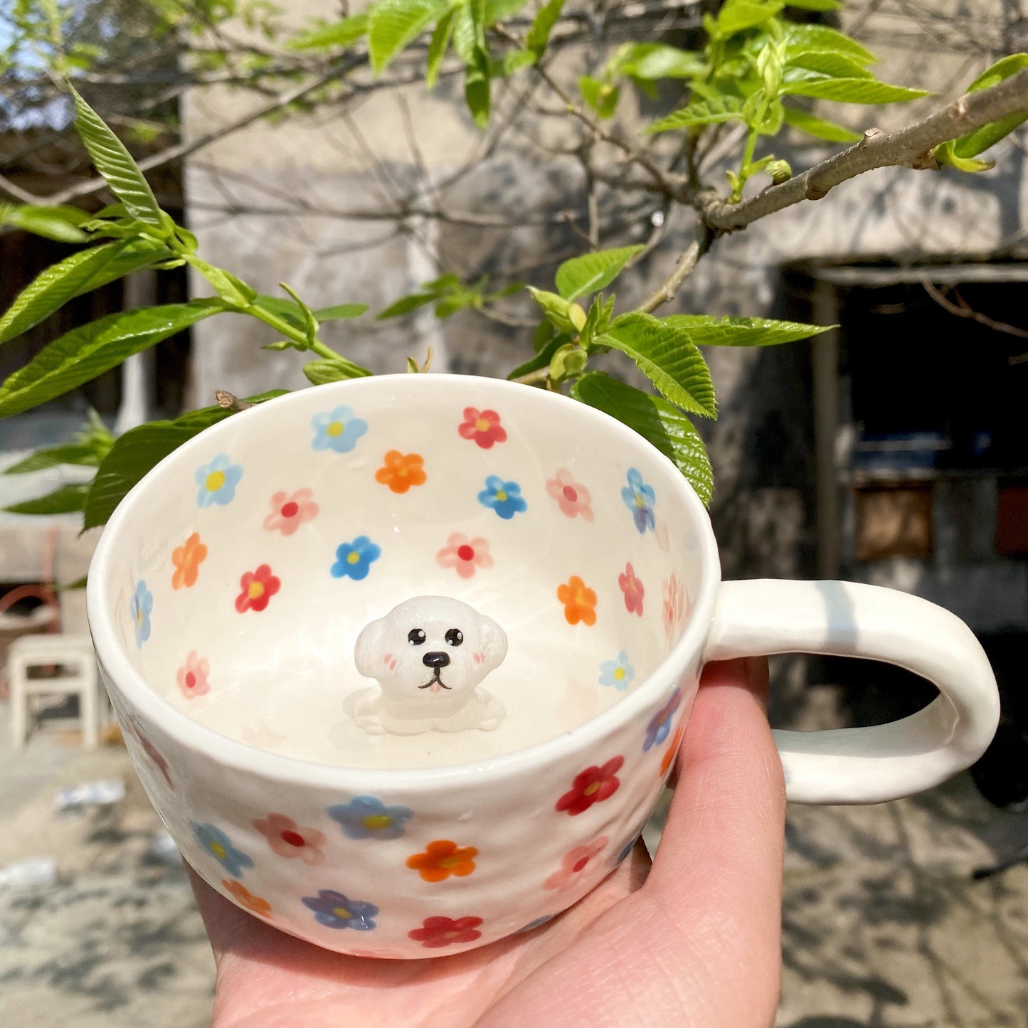 White ceramic mug with colorful hand-painted flowers and a 3D white puppy figurine inside, held outdoors near green leaves