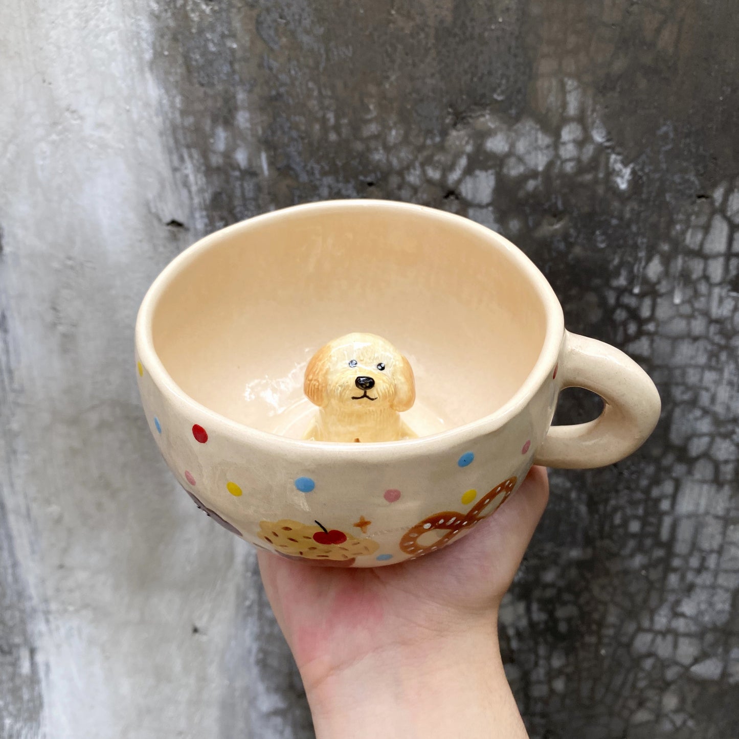 Cream-colored ceramic mug with colorful cookie and polka dot designs, featuring a 3D yellow puppy figurine inside, held by hand against a textured wall