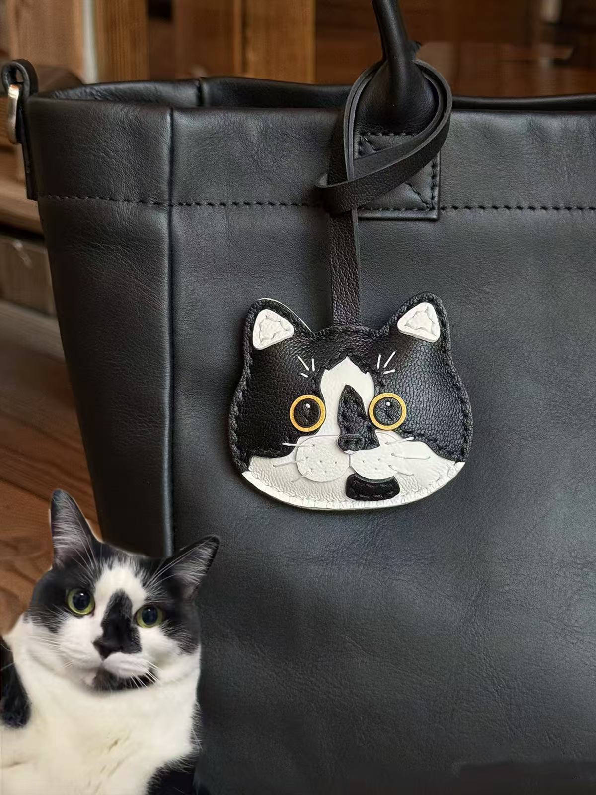 Realistic black and white cartoon cat charm hanging on a black leather bag, custom pet portrait made from goatskin leather
