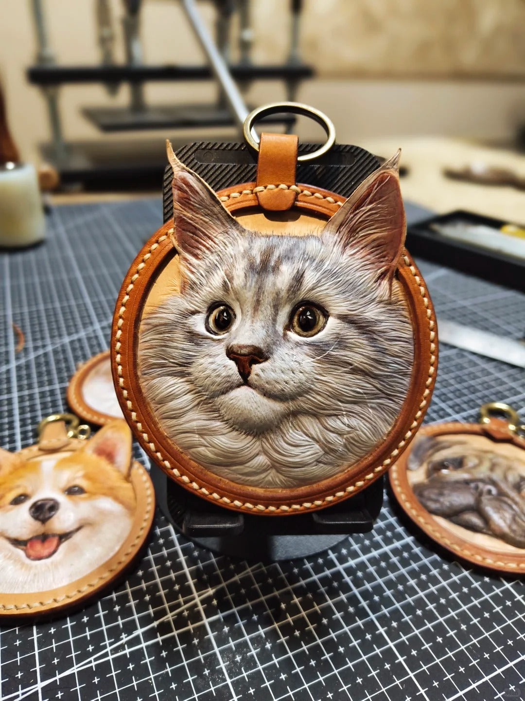 Handmade realistic leather hanging charm featuring a grey Maine Coon cat, carved and painted in 3D detail, shown on workbench with tools