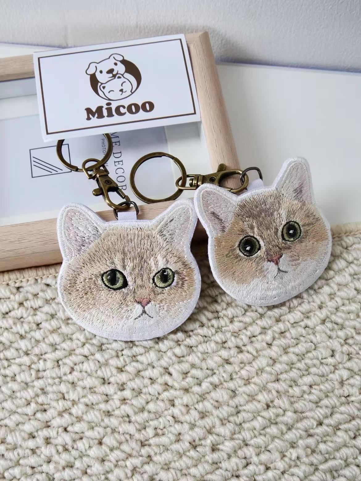 Pair of personalized embroidered round-face cat keychains with antique brass rings on beige fabric.