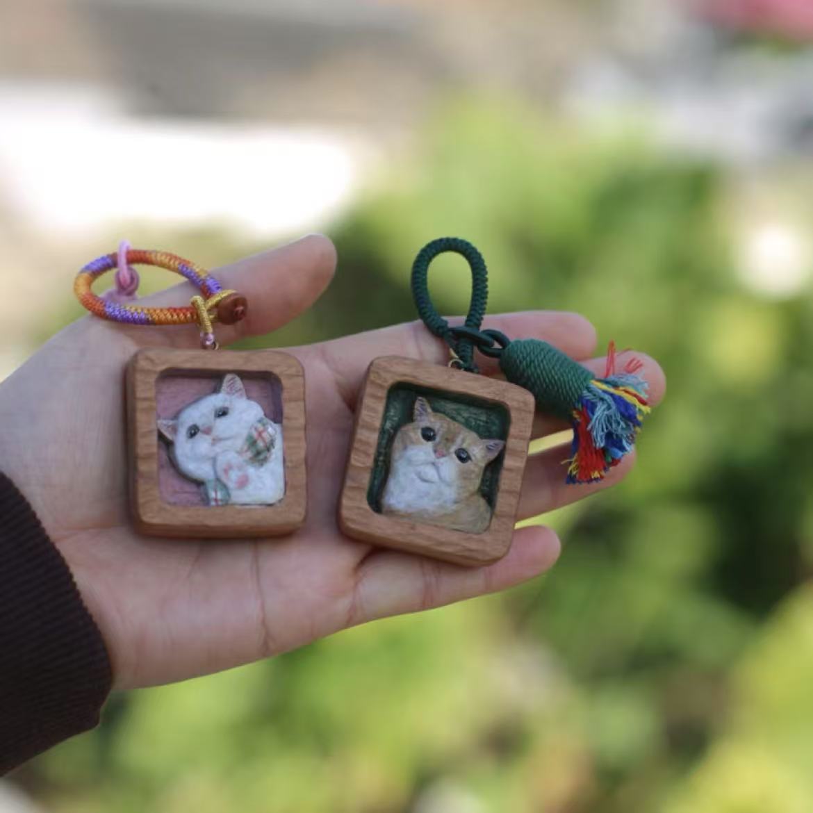 Two square wooden keychains, each featuring a hand-carved and painted cat portrait, decorated with colorful braided straps and tassels.