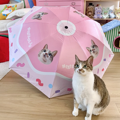 Cat sitting beside a pink custom umbrella printed with its portrait, indoor cozy setting.